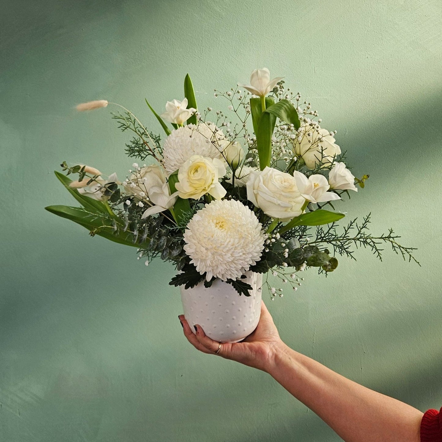 Hand holding a white ceramic vase filled with a bouquet of white flowers, including roses, chrysanthemums, and greenery, set against a muted green background.