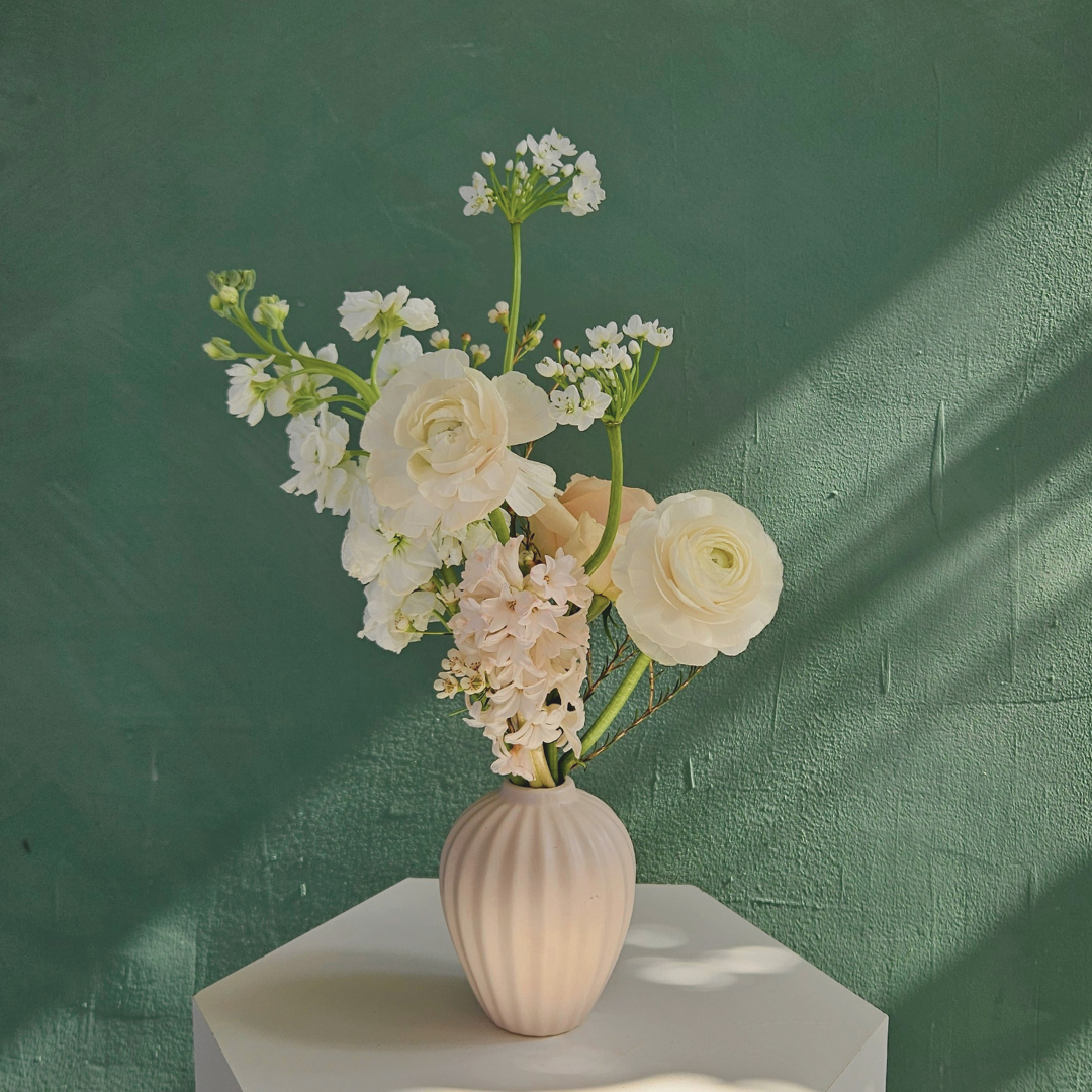 A ceramic white bud vase filled with white, cream and blush spring flowers , photographed against a green background