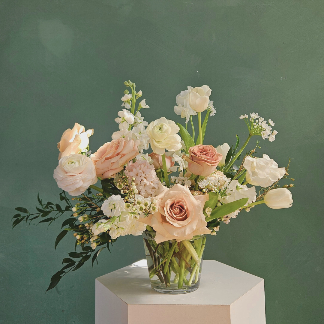 White, blush and tan spring flowers artfully arranged is a clear glass vase, photographed against a green background