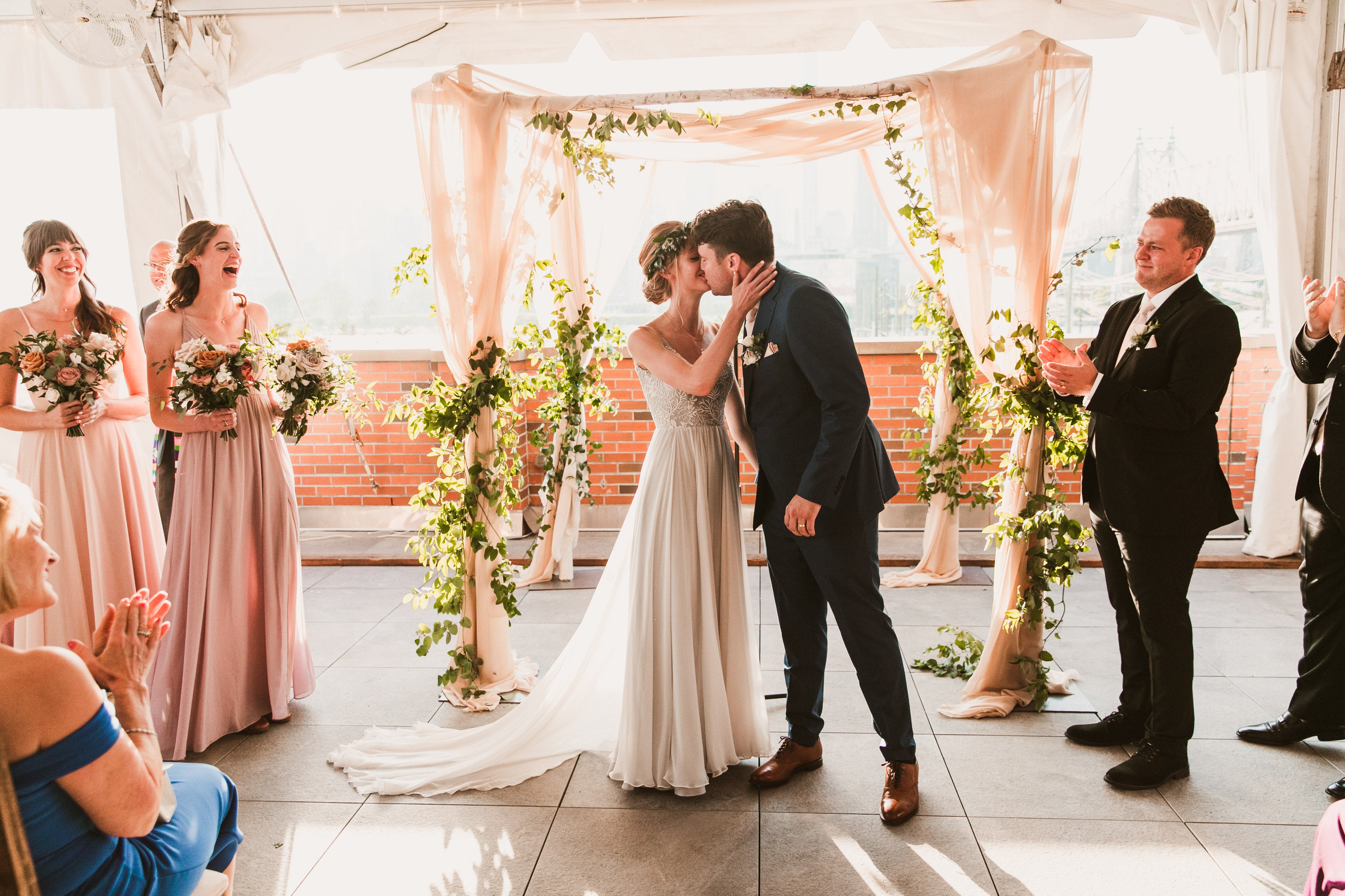 A joyful wedding ceremony as the bride and groom share their first kiss under a floral and fabric arch, surrounded by bridesmaids, groomsmen, and guests applauding in celebration.