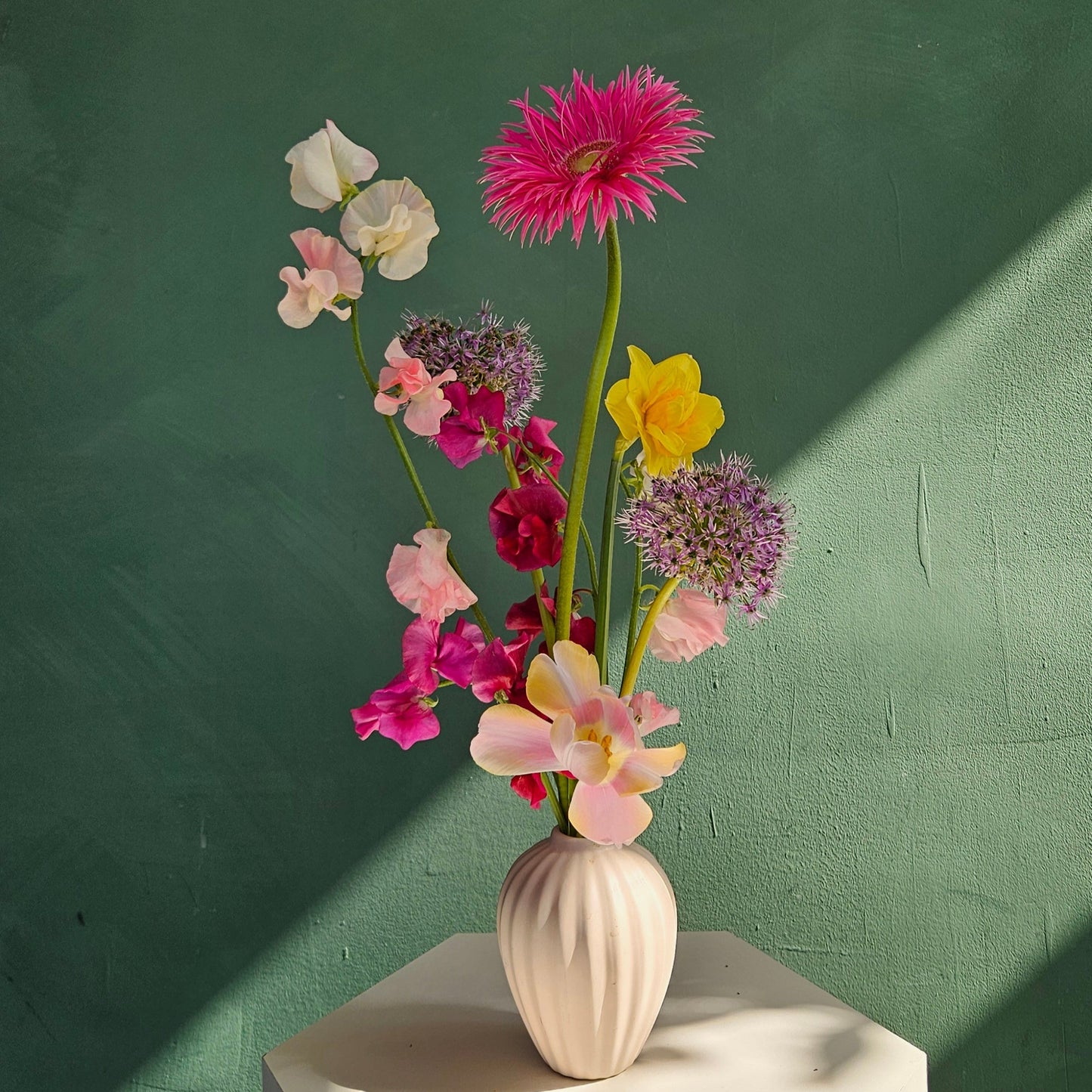 A white ceramic bud vase filled with colorful spring flowers, photographed against a green background