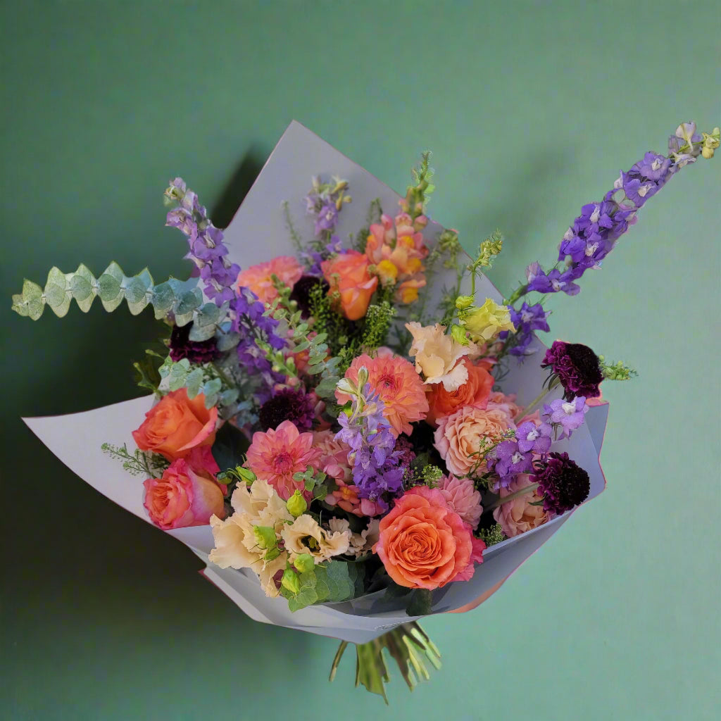 A vibrant mixed floral bouquet featuring orange roses, pink dahlias, purple delphiniums, and green eucalyptus, wrapped in light paper. Perfect for special occasions or gifting.