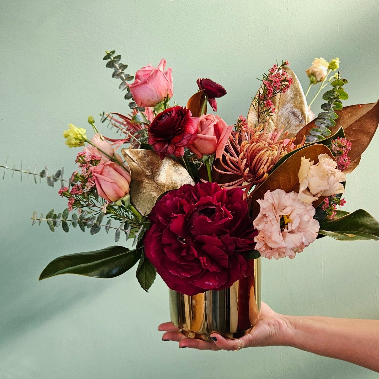 Hand holding a gold vase filled with an arrangement of vibrant flowers, including deep red and pink roses, burgundy ranunculus, chrysanthemums, blush lisianthus, eucalyptus, and golden accent leaves, against a muted green background.