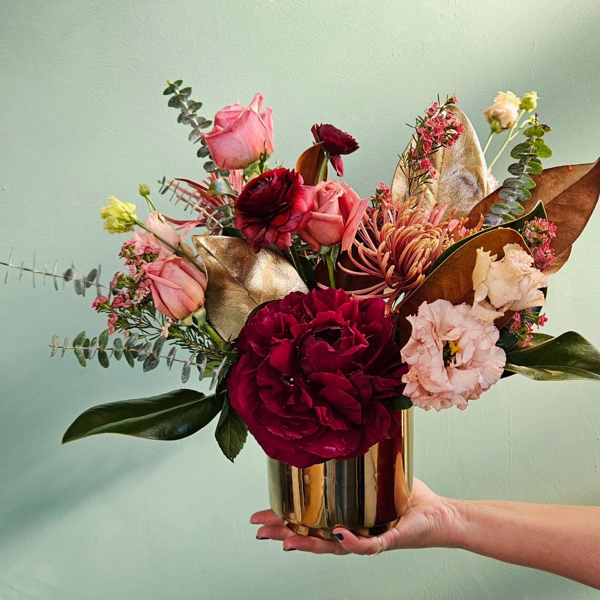 Hand holding a gold vase filled with an arrangement of vibrant flowers, including deep red and pink roses, burgundy ranunculus, chrysanthemums, blush lisianthus, eucalyptus, and golden accent leaves, against a muted green background.