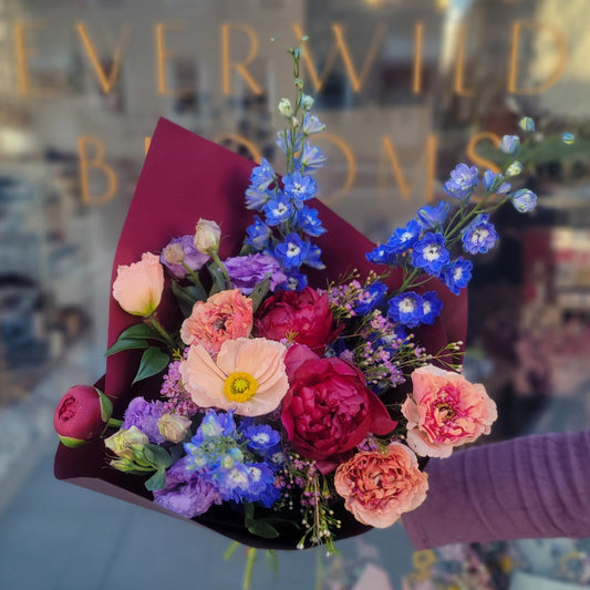 A vibrant bouquet of flowers including roses, peonies, and various other seasonal blooms, wrapped in pink paper.