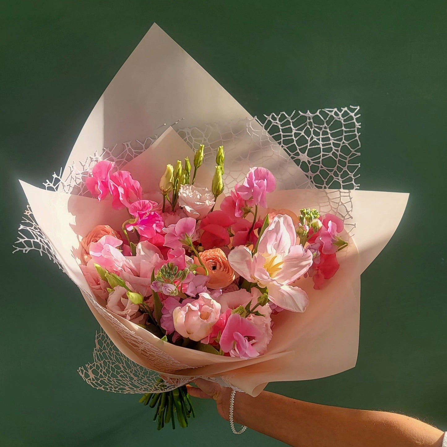 A stunning bouquet of pink spring flowers, including sweet peas, ranunculus, and lisianthus, elegantly wrapped in light peach paper with decorative lace accents, held against a dark green background.