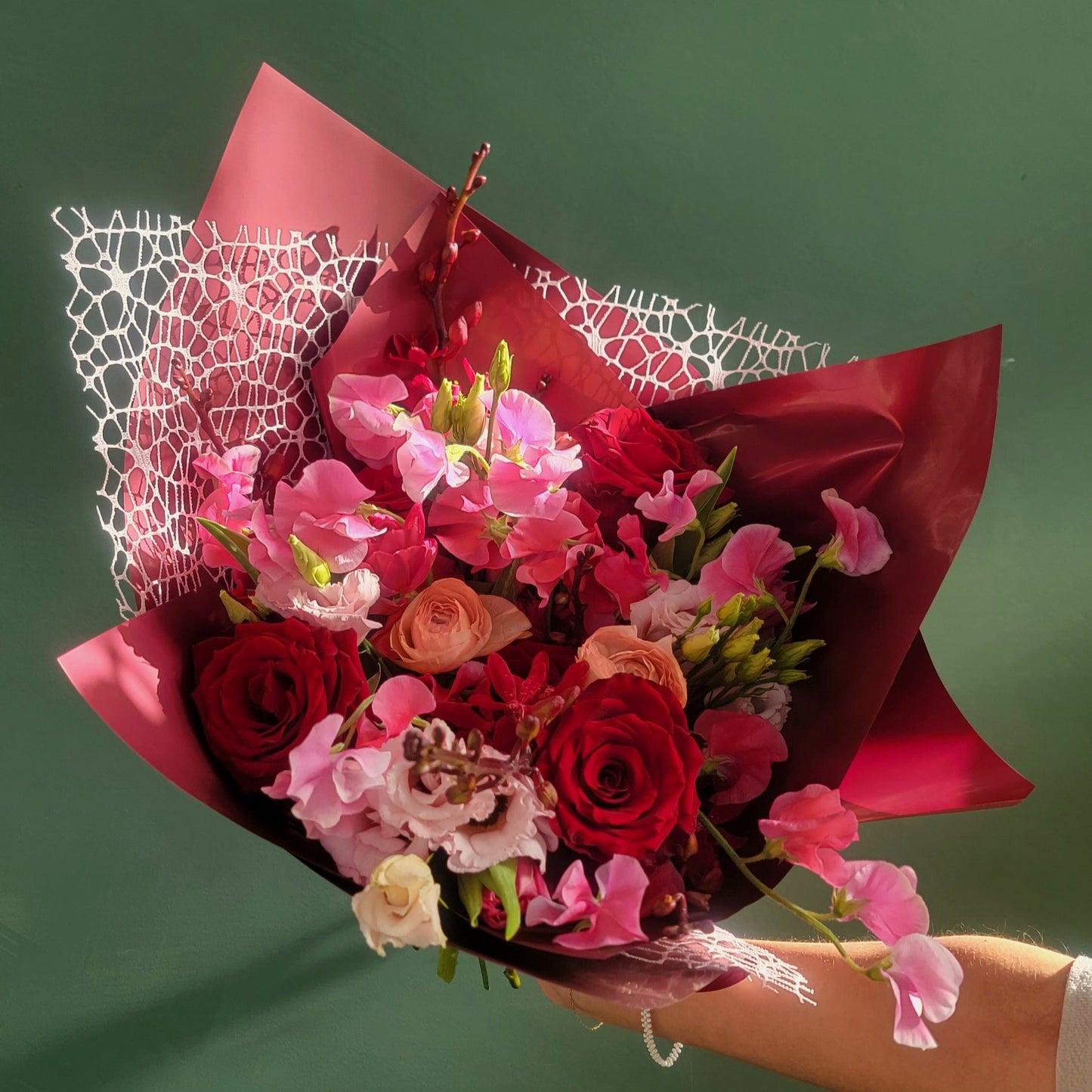 A romantic bouquet featuring deep red roses, soft pink sweet peas, blush ranunculus, and delicate lisianthus, wrapped in rich red paper with a lace accent, held against a green background.