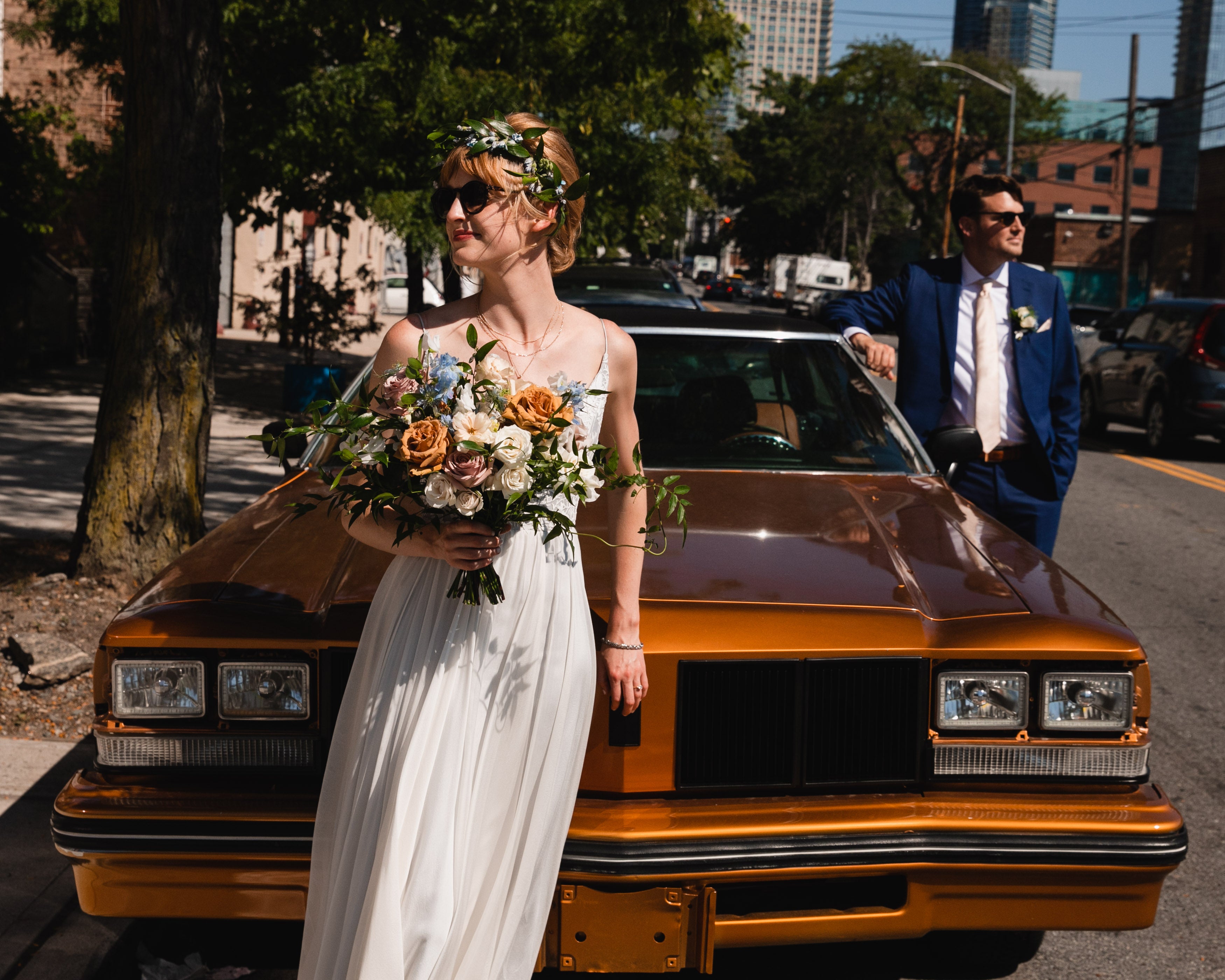 A stylish bride holding a rustic floral bouquet, standing against a vintage bronze car, with the groom leaning casually nearby in a navy suit.