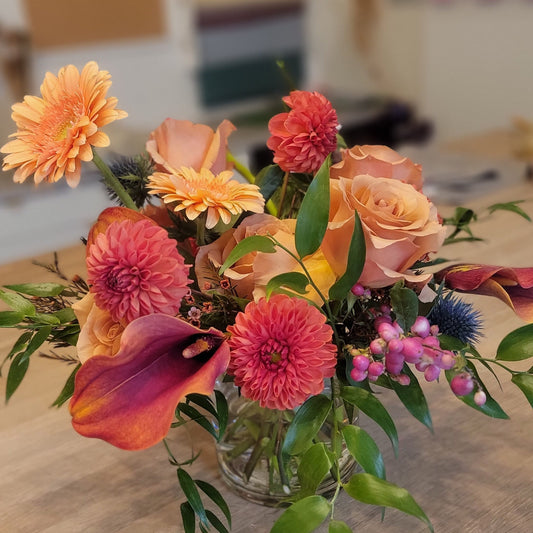 A lush floral arrangement in warm autumn hues with peach roses, orange dahlias, calla lilies, and hypericum berries, displayed in a glass vase.