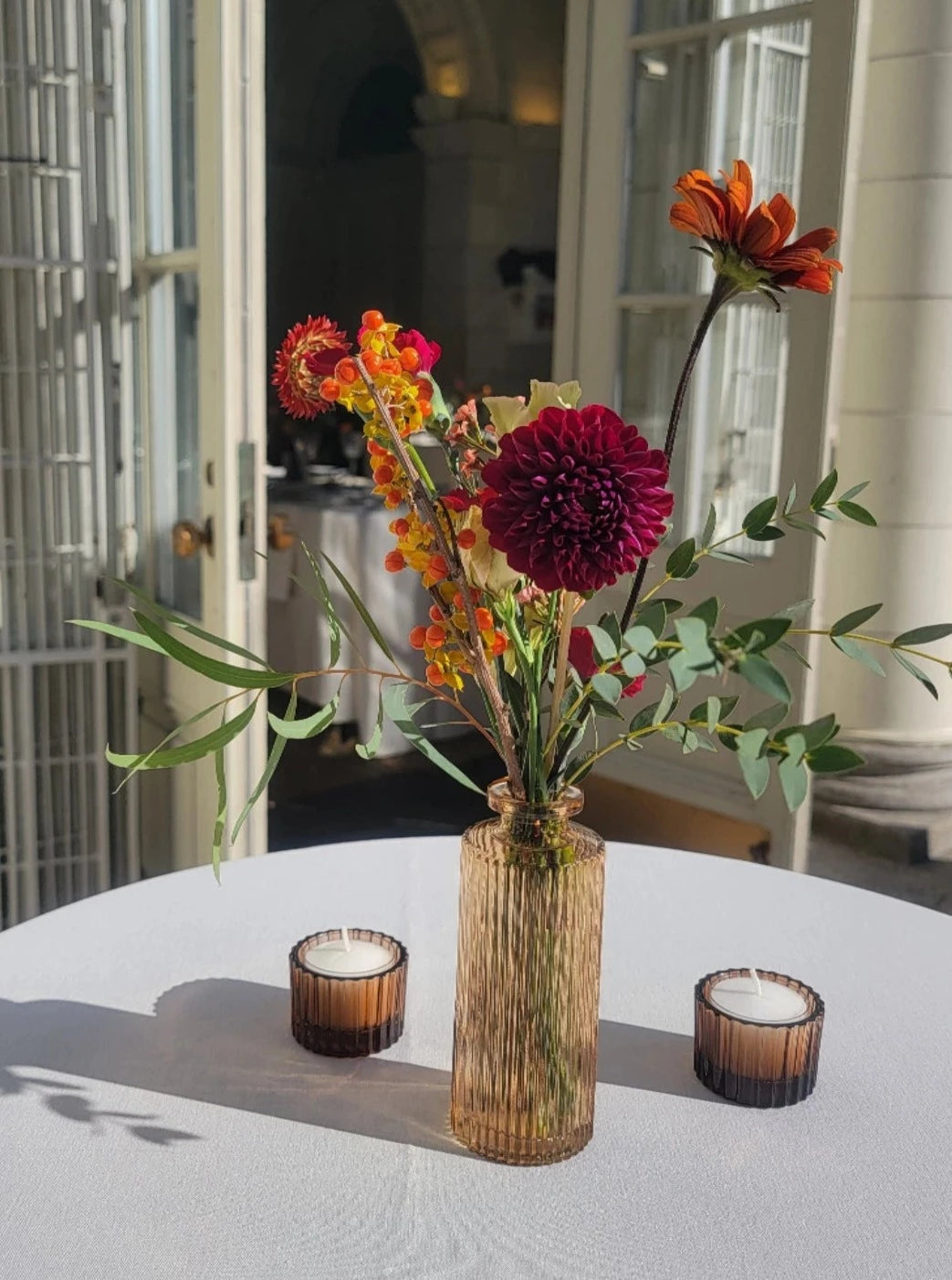 A tall amber glass vase with a floral arrangement featuring deep burgundy dahlia, orange zinnia, yellow snapdragons, and eucalyptus, styled on a white table with two amber candle holders.