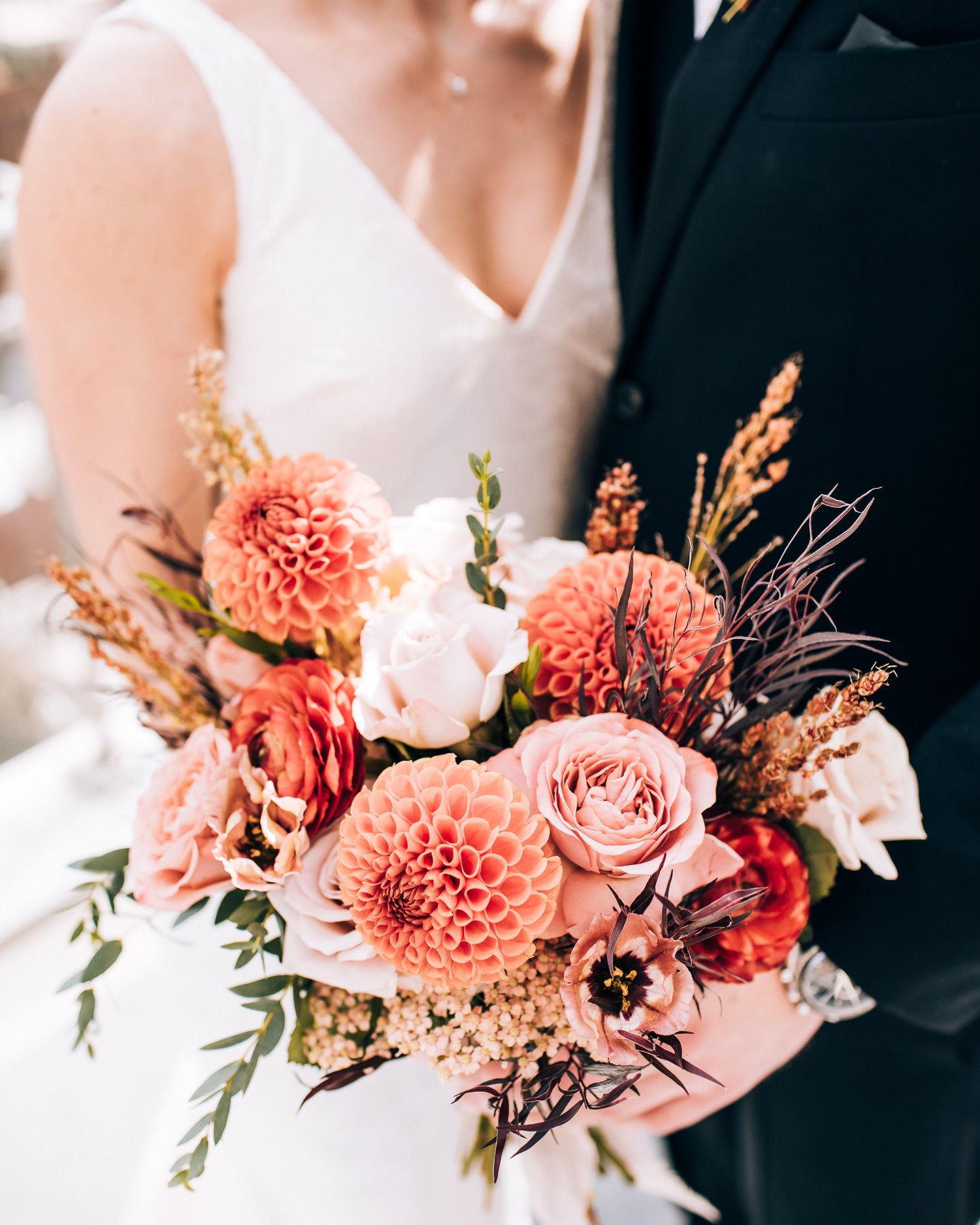 A close-up of a bridal bouquet featuring peach dahlias, soft pink roses, ranunculus, and seasonal greenery, held by the bride and groom in elegant wedding attire.