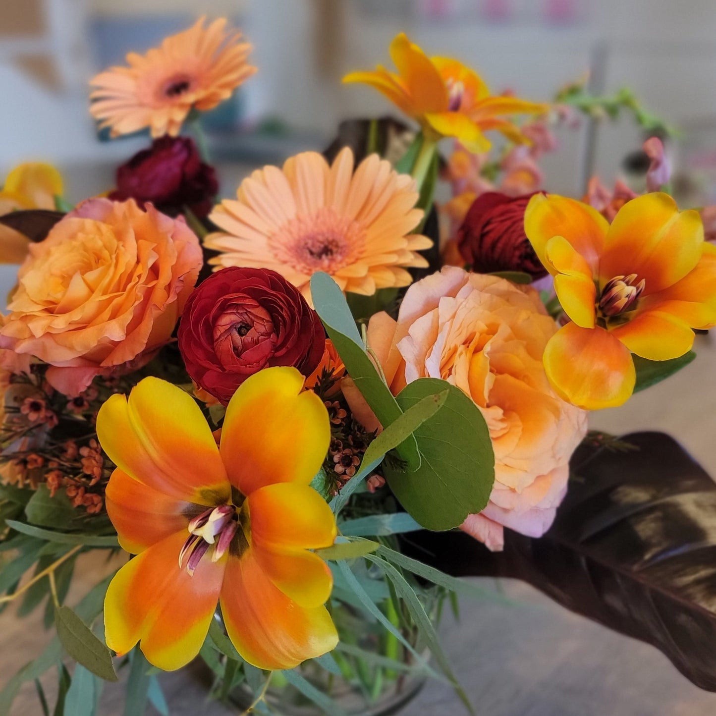 A cheerful floral arrangement with bright orange tulips, yellow gerbera daisies, peach roses, and burgundy ranunculus in a glass vase.