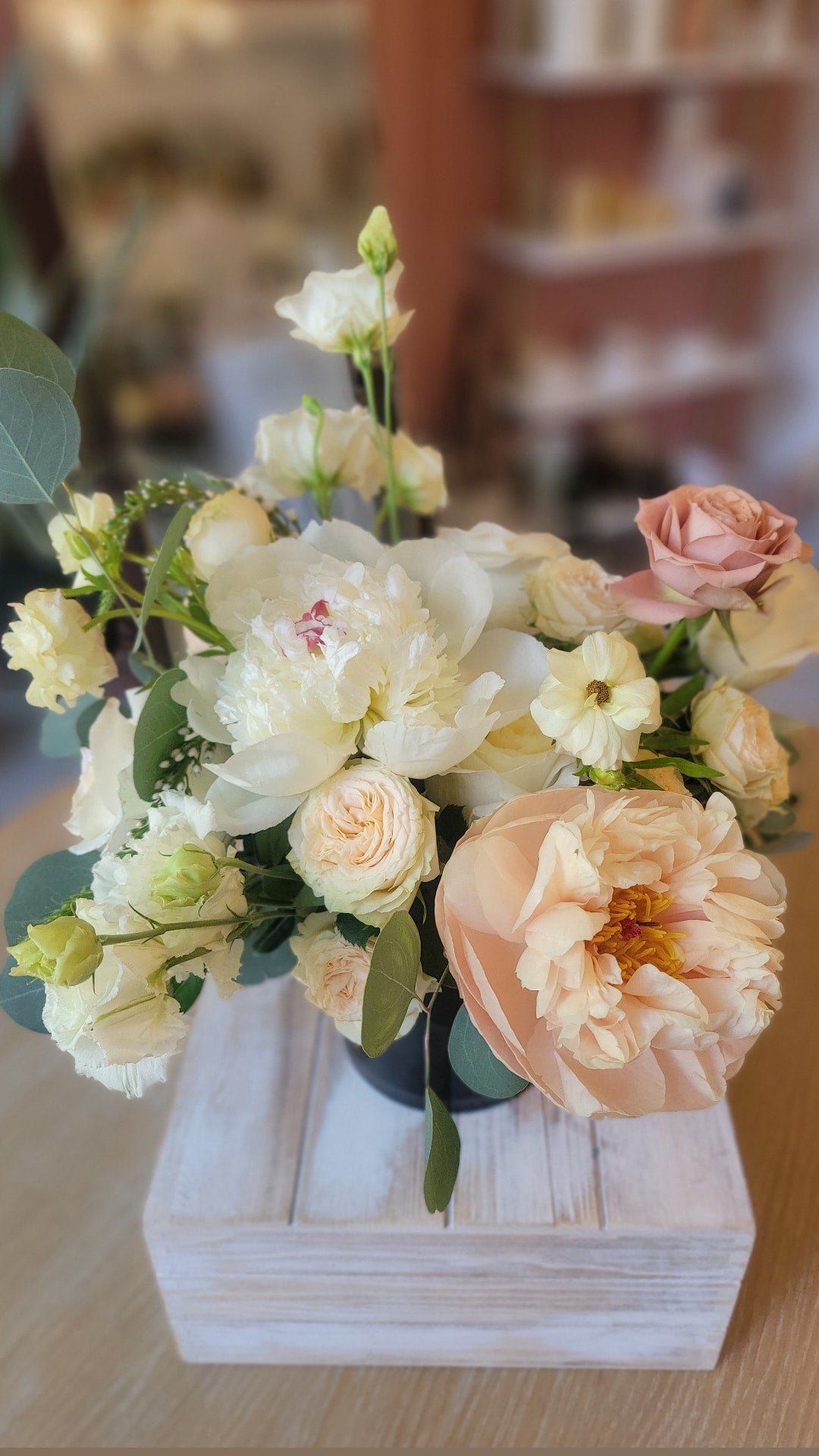 A soft and elegant floral arrangement with white peonies, peach garden roses, and lisianthus in a black vase, placed on a wooden box.