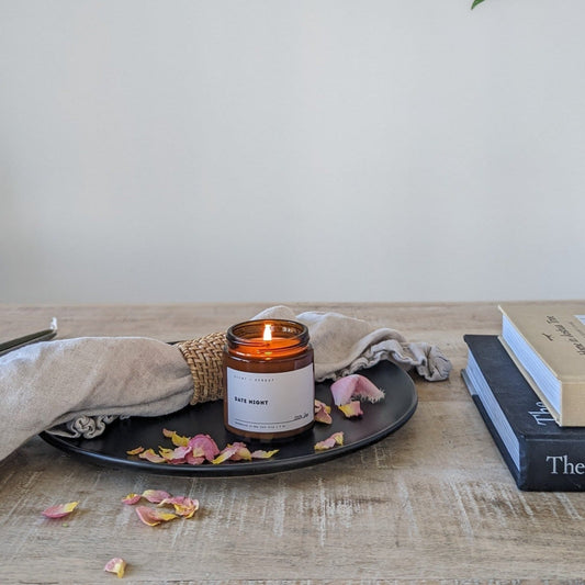 A lit amber jar candle labeled "Date Night" with a minimal design, surrounded by soft fabric, dried rose petals, and warm ambiance.
