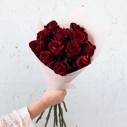 A stunning bouquet of deep red roses, elegantly arranged and wrapped in soft pink paper, displayed against a white background.