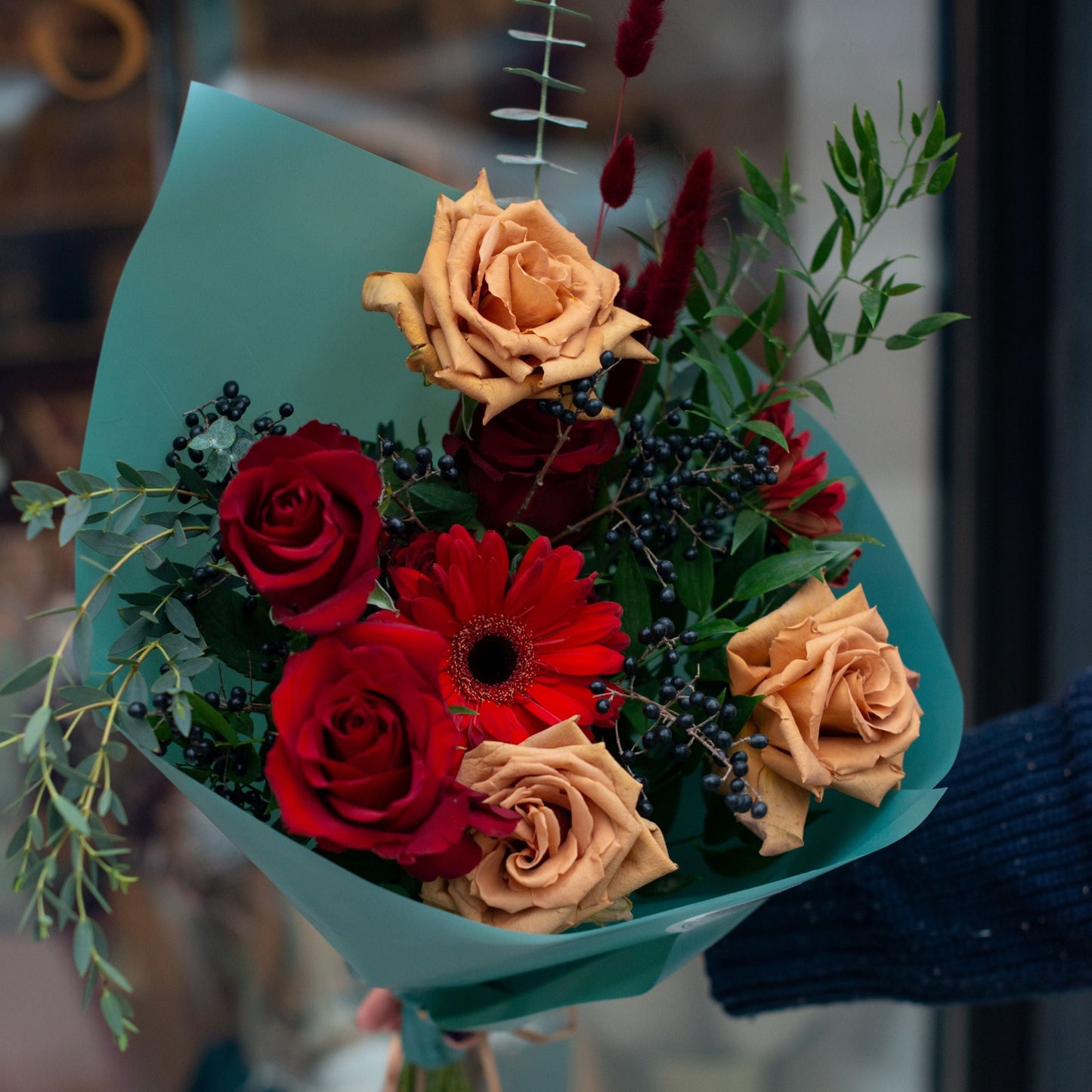 A sophisticated bouquet with deep red roses, orange-gold roses, red gerbera daisies, black berries, and greenery, wrapped in a soft teal paper for a striking presentation.