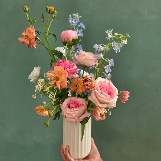 Whimsical garden-style bouquet with pink roses, ranunculus, and delphiniums in a white ceramic vase, held by hand.