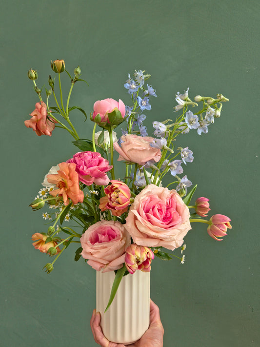 Whimsical garden-style bouquet with pink roses, ranunculus, and delphiniums in a white ceramic vase, held by hand.