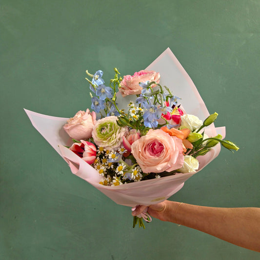 Fresh spring bouquet with pink roses, ranunculus, tulips, chamomile, and delphinium wrapped in pastel pink paper.