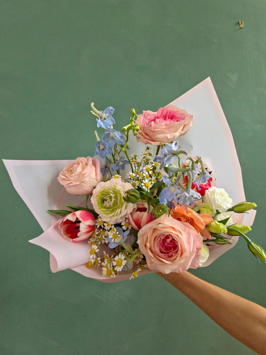 Fresh spring bouquet with pink roses, ranunculus, tulips, chamomile, and delphinium wrapped in pastel pink paper.