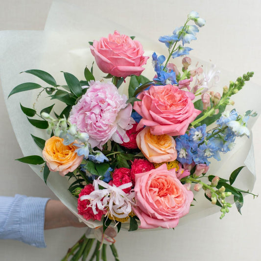 Colorful spring bouquet with garden roses, peonies, delphiniums, and seasonal blooms wrapped in white paper.