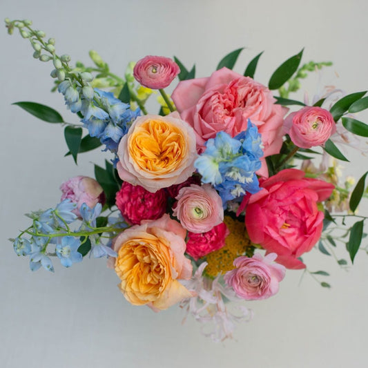Colorful spring bouquet with garden roses, peonies, delphiniums, and seasonal blooms wrapped in white paper.