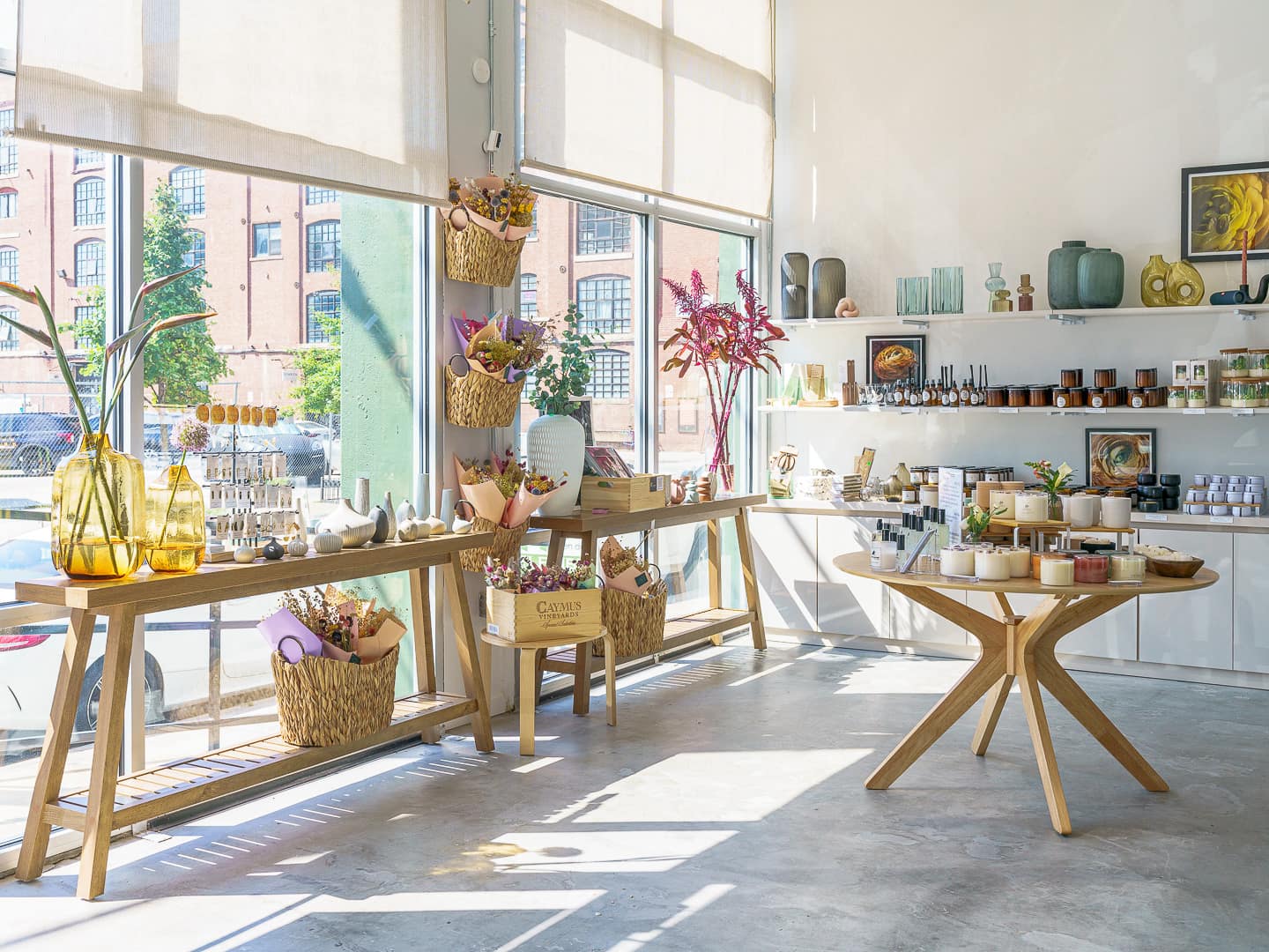 A bright and inviting floral studio showcasing handcrafted bouquets, elegant vases, candles, and artisanal goods, illuminated by natural sunlight streaming through large windows.