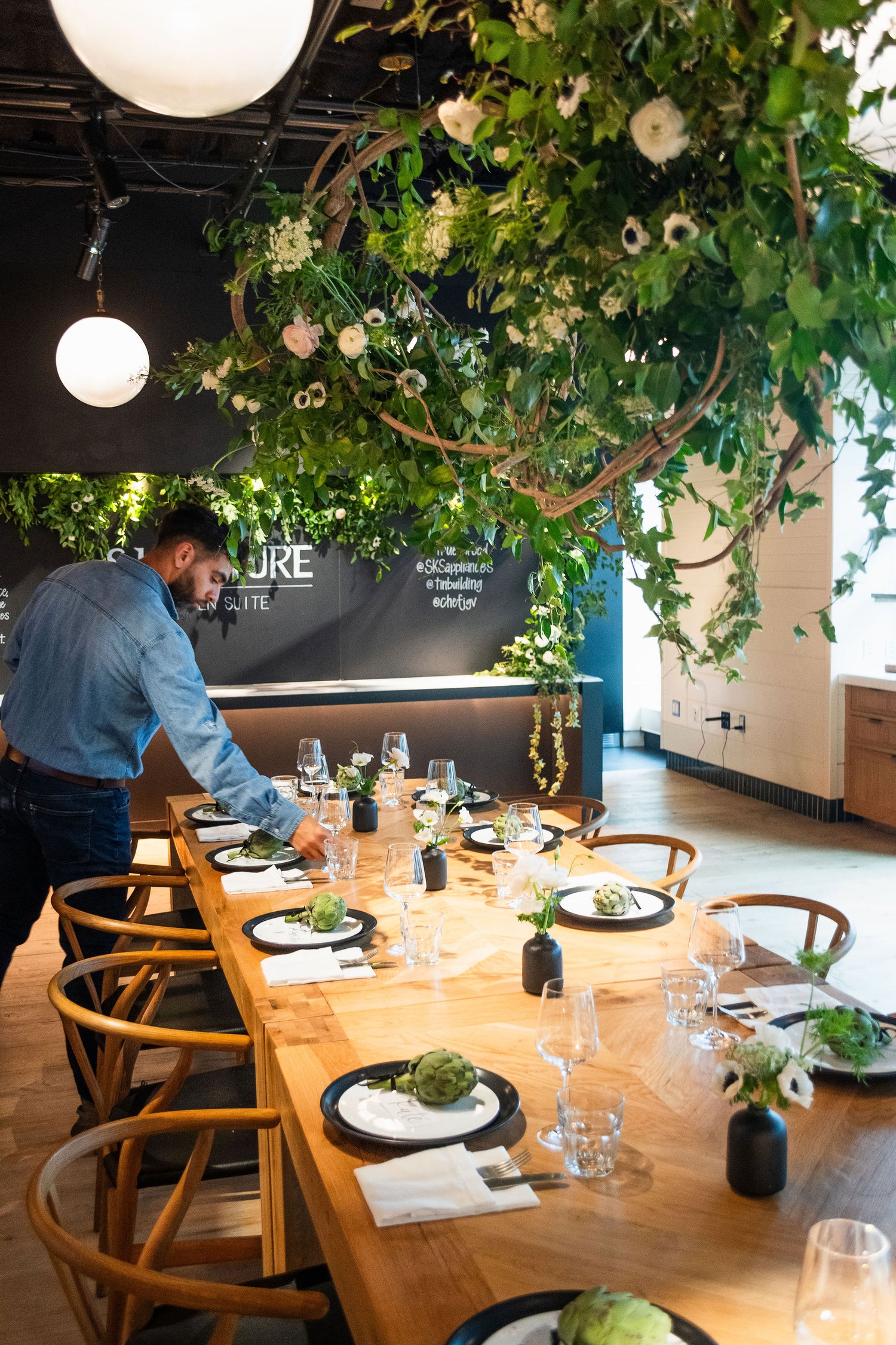 A stylish dining table set for an event, adorned with black vases, greenery, and artichoke accents, beneath a lush overhead greenery installation. A person adjusts the table setting in a modern space.