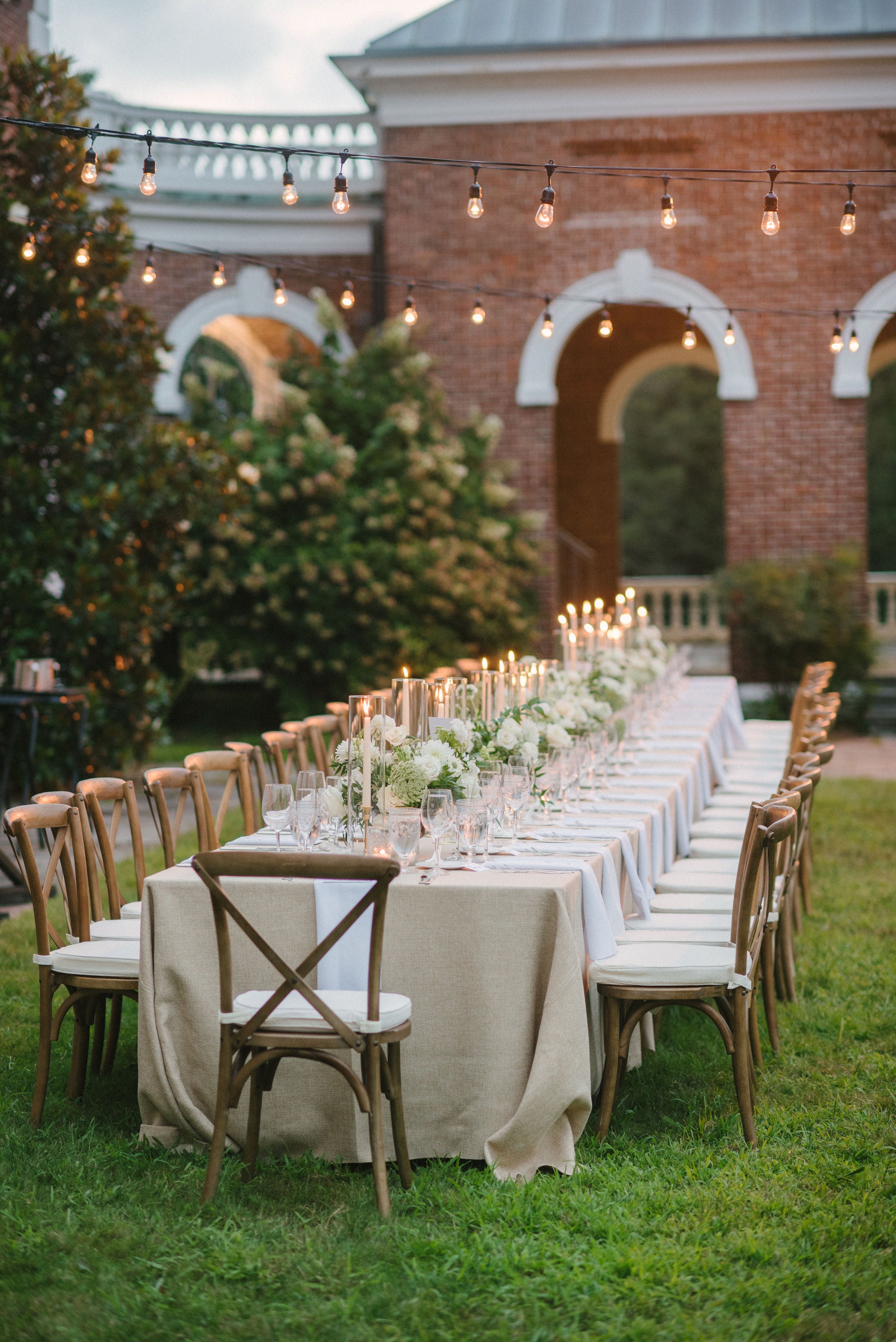 A beautifully set outdoor wedding reception table adorned with white florals, candles, and string lights, surrounded by lush greenery and a charming brick venue.