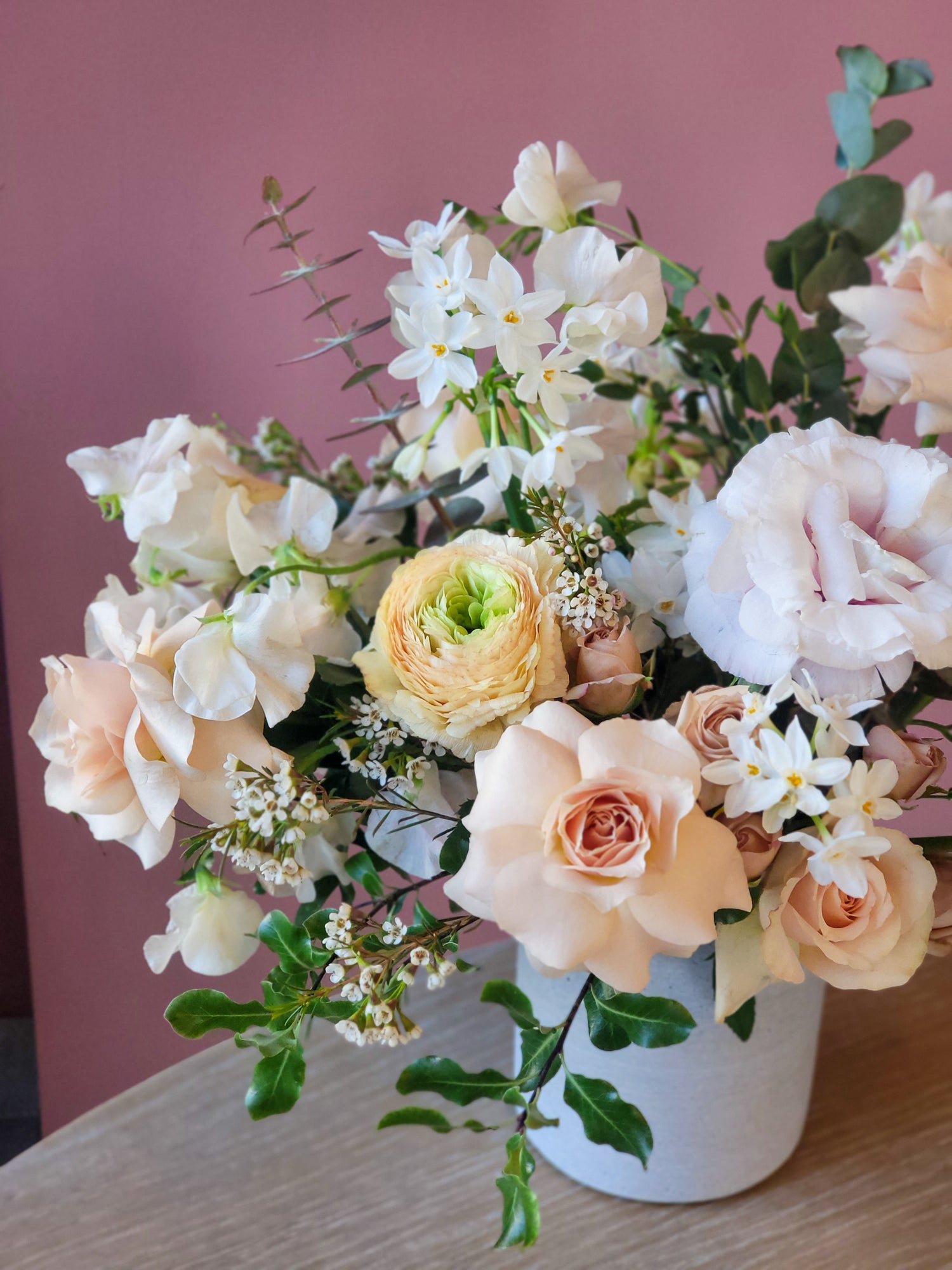 Elegant floral arrangement featuring pastel roses, white blossoms, and greenery in a white vase against a soft pink background.