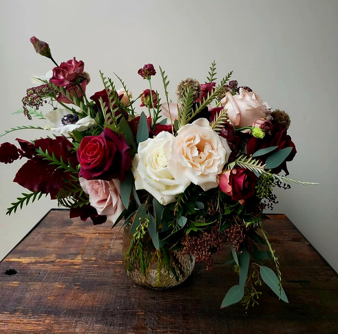 A dramatic arrangement featuring burgundy and cream roses, ferns, and eucalyptus, styled in a clear textured vase on a wooden table.