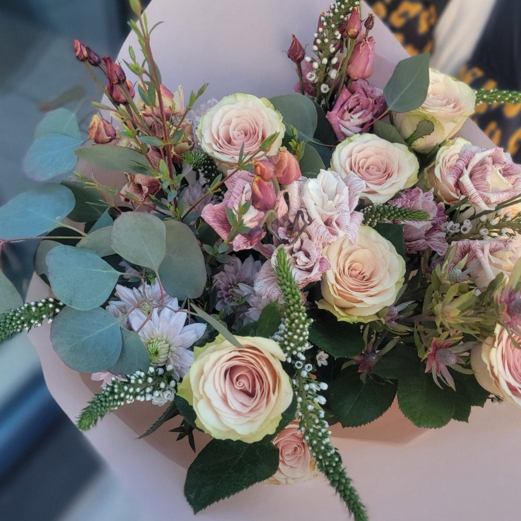 A delicate bouquet featuring pale pink roses, mauve lisianthus, eucalyptus leaves, and white veronica flowers, wrapped in soft blush paper for an elegant touch.