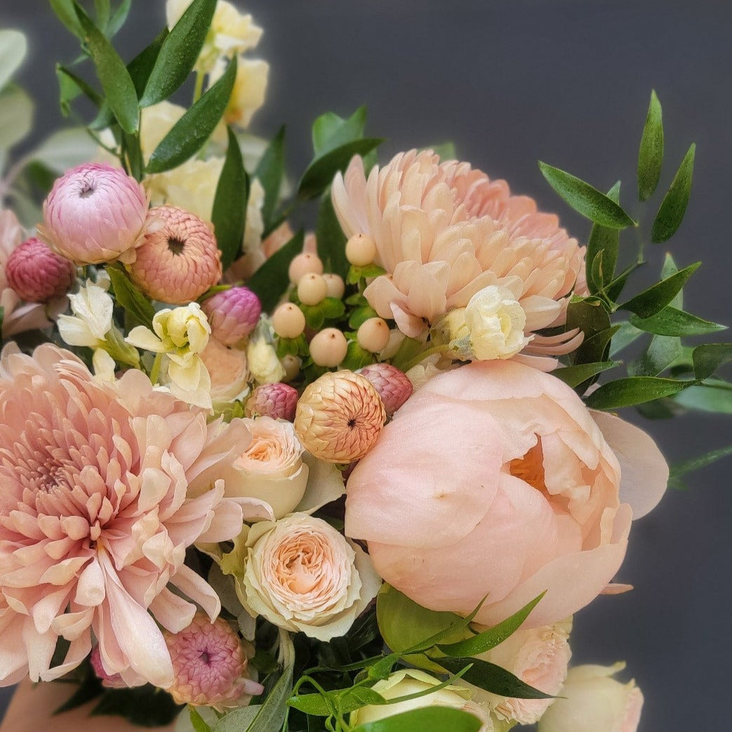 A delicate pastel floral arrangement with blush pink peonies, cream roses, pale chrysanthemums, and hypericum berries, styled with soft greenery.