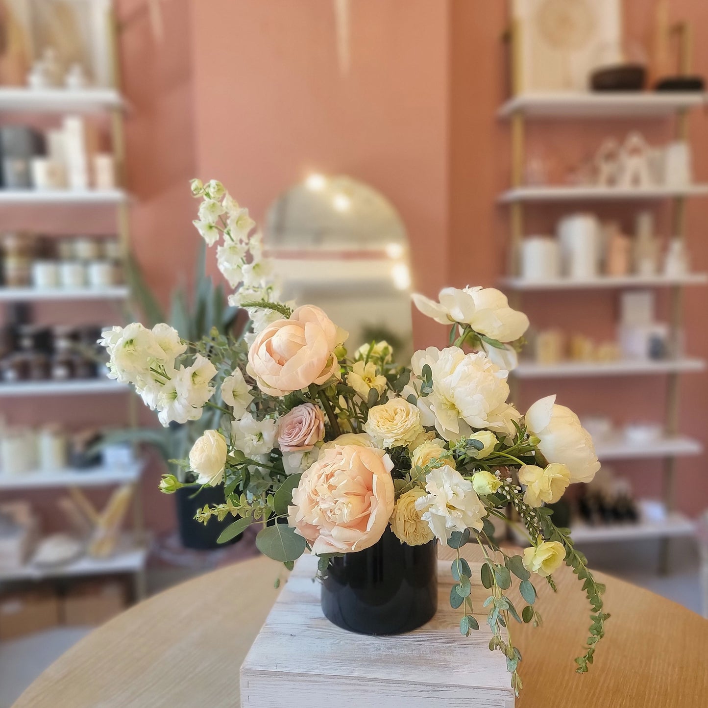 A delicate display with cream and peach roses, white ranunculus, and snapdragons arranged in a black vase, set against a soft pink background.