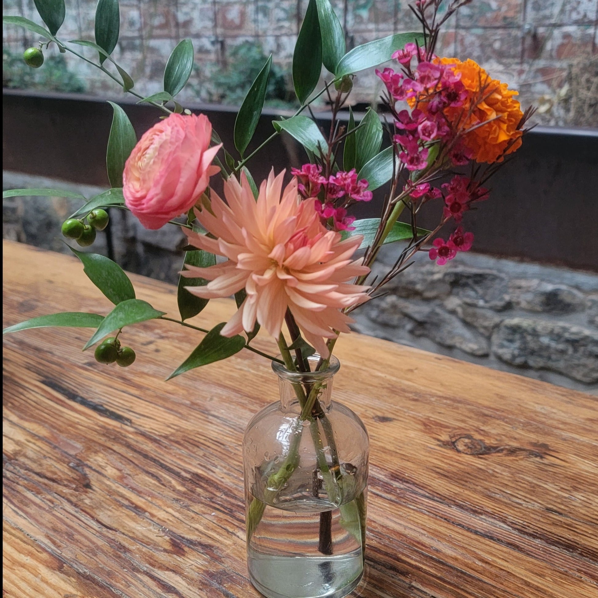 A small floral arrangement in a clear glass bud vase featuring a soft pink dahlia, pink ranunculus, orange marigold, and wax flowers, set on a rustic wooden table.