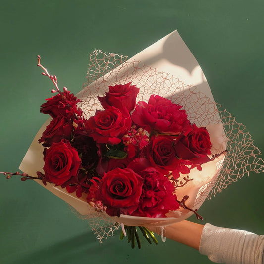 A stunning bouquet of crimson red roses and lush peonies, accented with delicate red blossoms and wrapped in elegant white paper with lace detail, held against a green background.