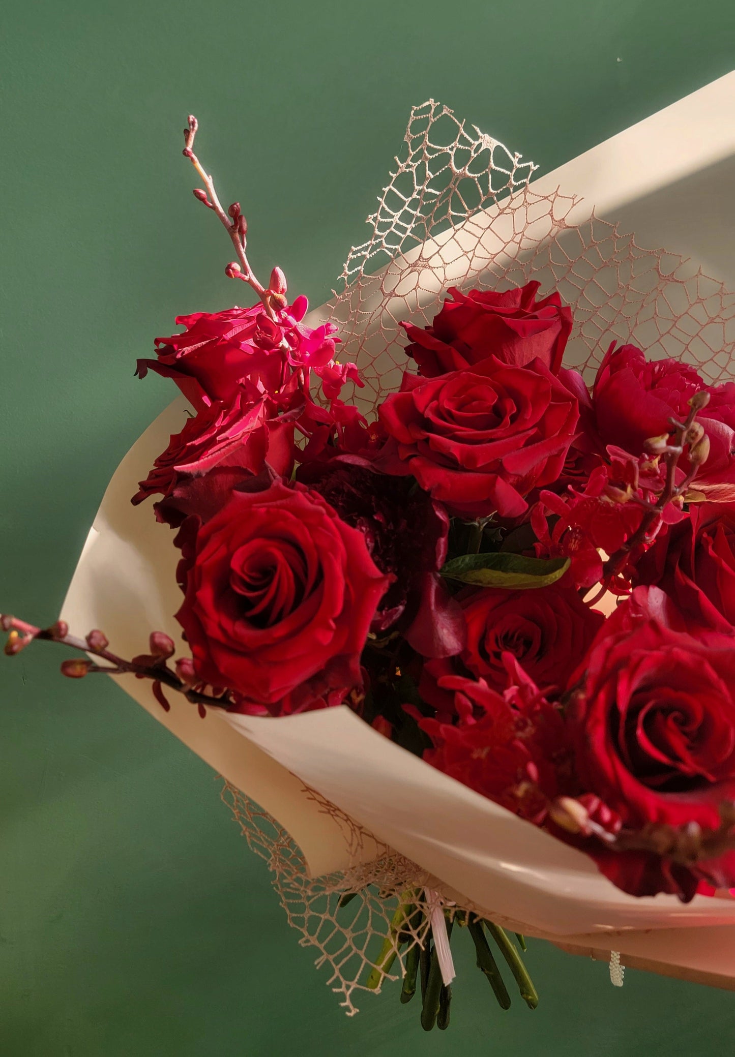 A stunning bouquet of crimson red roses and lush peonies, accented with delicate red blossoms and wrapped in elegant white paper with lace detail, held against a green background.