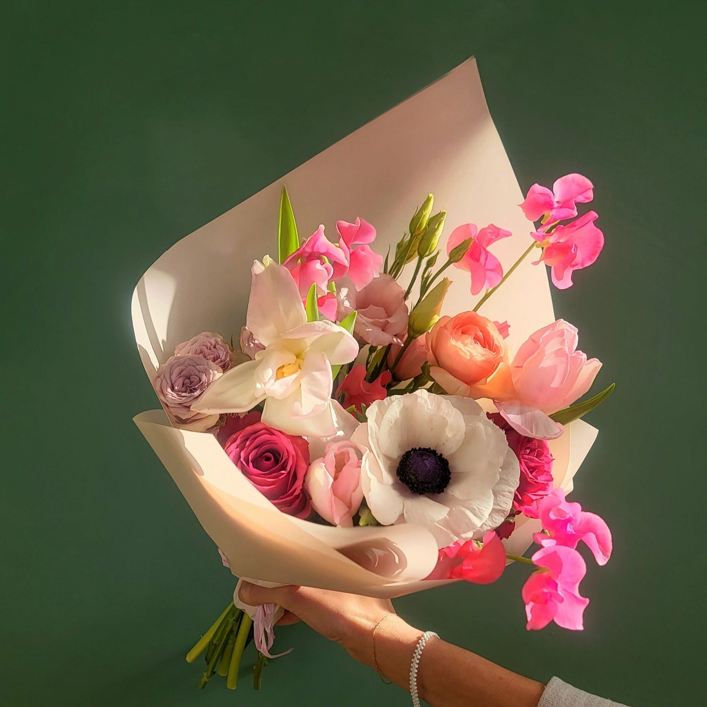 A radiant bouquet of pastel flowers including white anemones, blush roses, and pink sweet peas, elegantly wrapped in cream paper, held against a soft green backdrop.