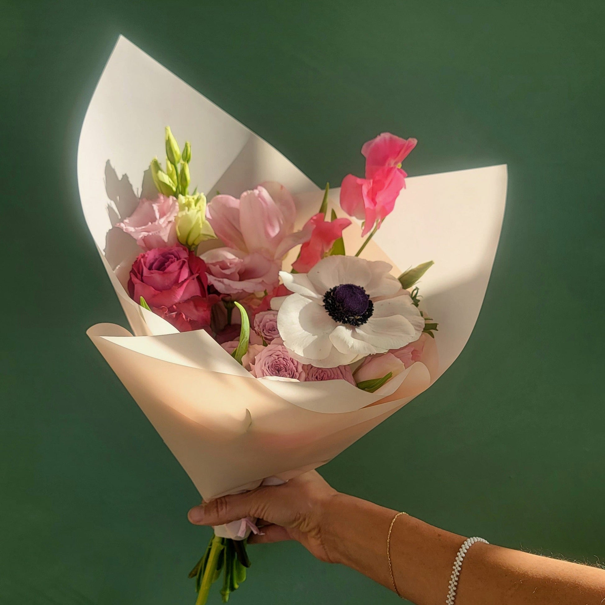 A delicate bouquet featuring white anemones, soft pink tulips, lavender roses, and sweet peas, wrapped in cream paper, held against a green background.