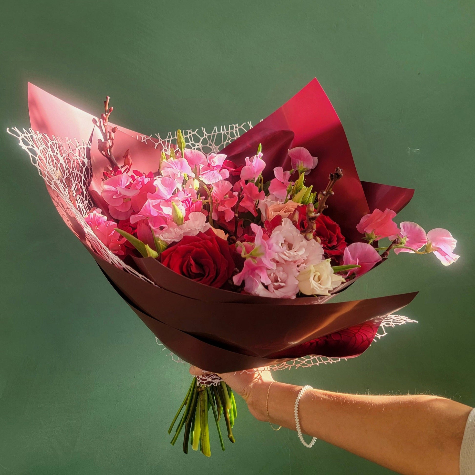 A romantic bouquet featuring deep red roses, soft pink sweet peas, blush ranunculus, and delicate lisianthus, wrapped in rich red paper with a lace accent, held against a green background.