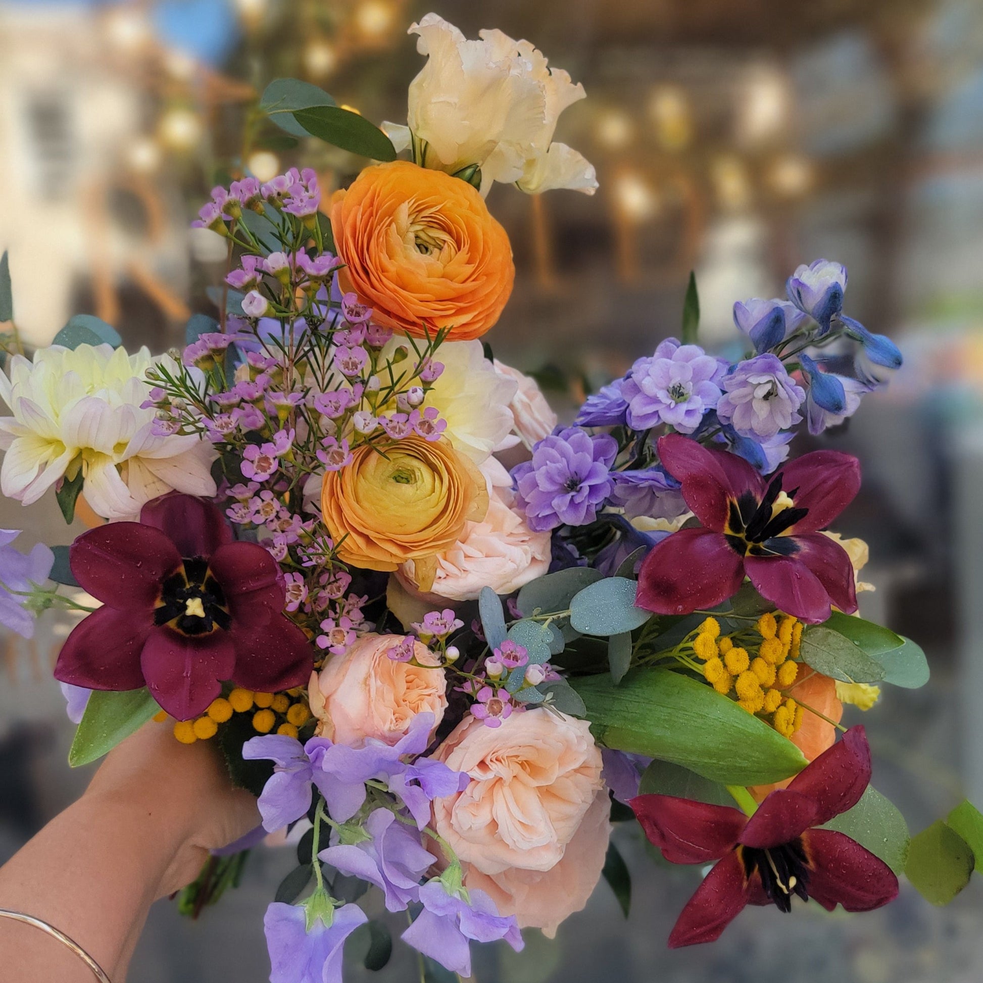 A cheerful bouquet featuring orange ranunculus, deep red tulips, purple delphiniums, peach roses, and wax flowers with lush greenery, perfect for a colorful celebration.