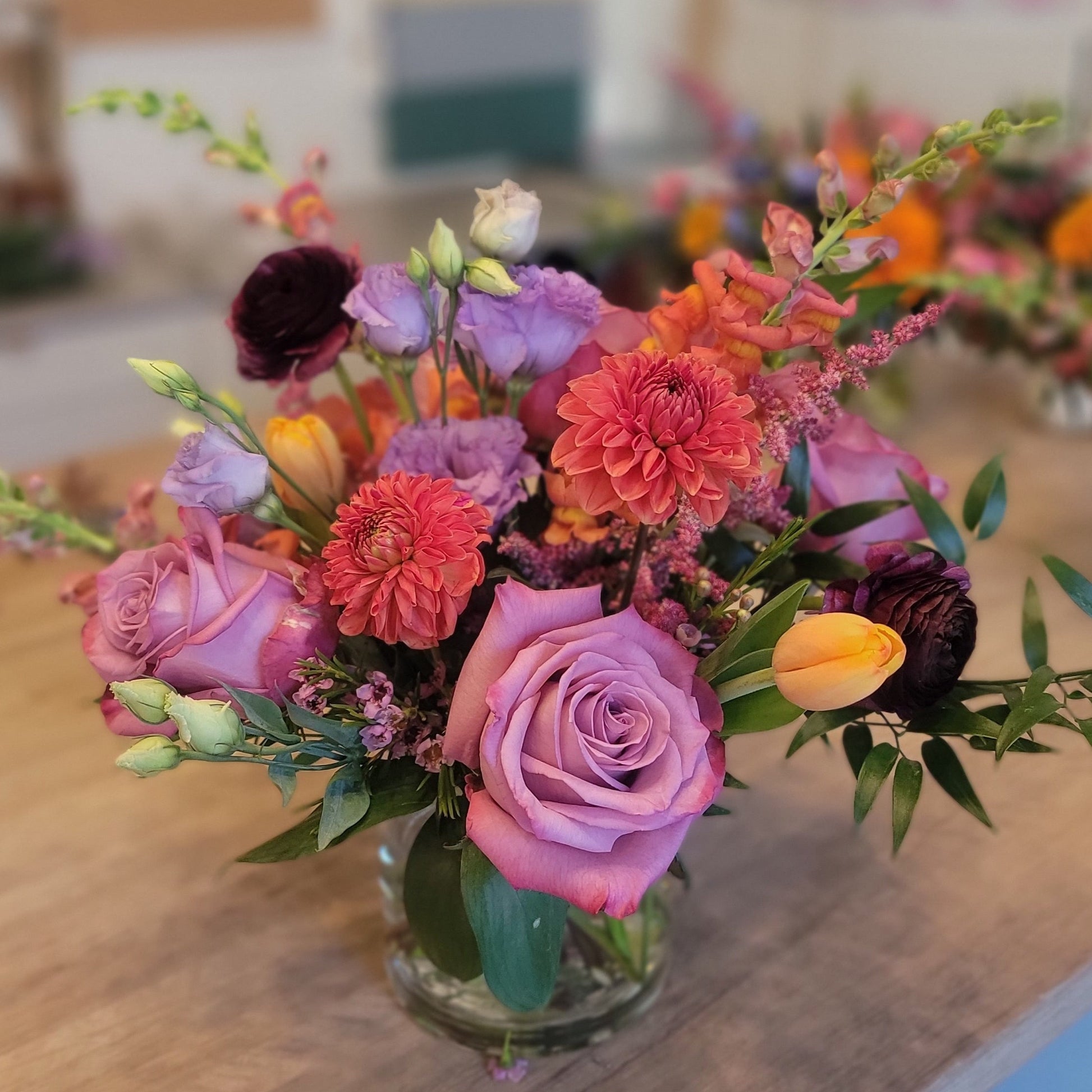 A vibrant centerpiece with purple roses, orange zinnias, tulips, and lisianthus in a glass vase, perfect for special occasions.