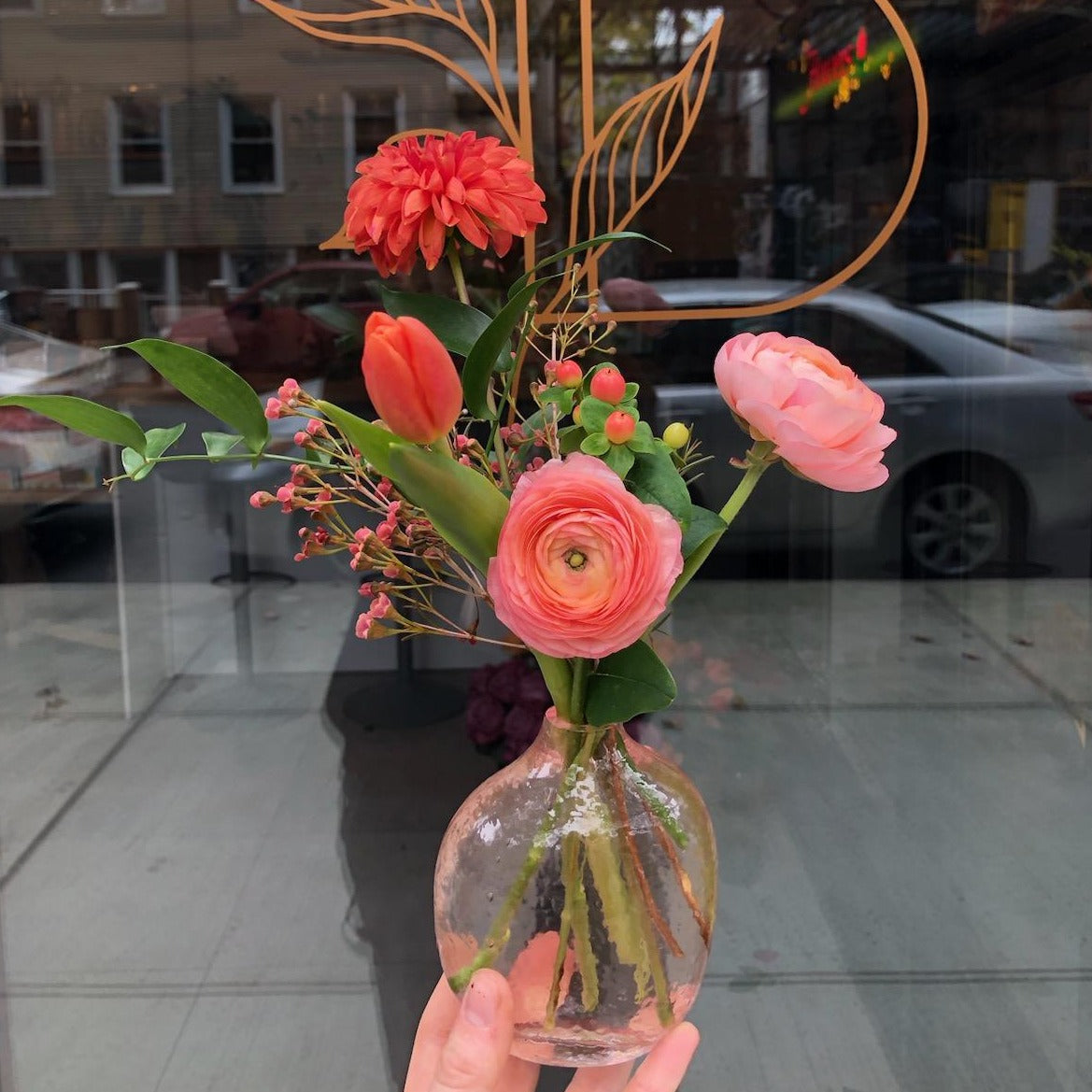 A delicate floral arrangement in a clear glass bud vase featuring pink ranunculus, orange dahlia, tulip, and hypericum berries, perfect for small spaces or gift accents.