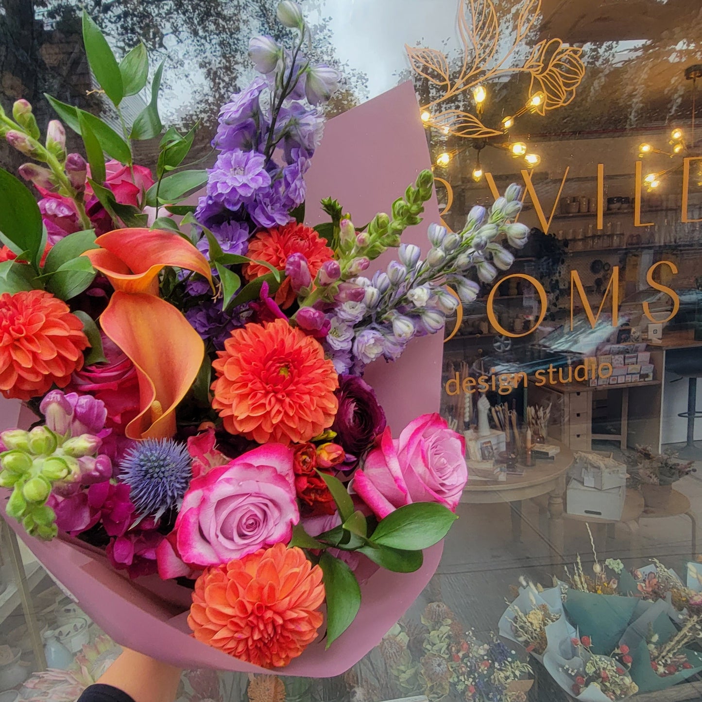 A bold and colorful bouquet with bright orange dahlias, pink roses, purple delphiniums, orange calla lilies, and thistle, wrapped in vibrant pink paper, ideal for celebrations.