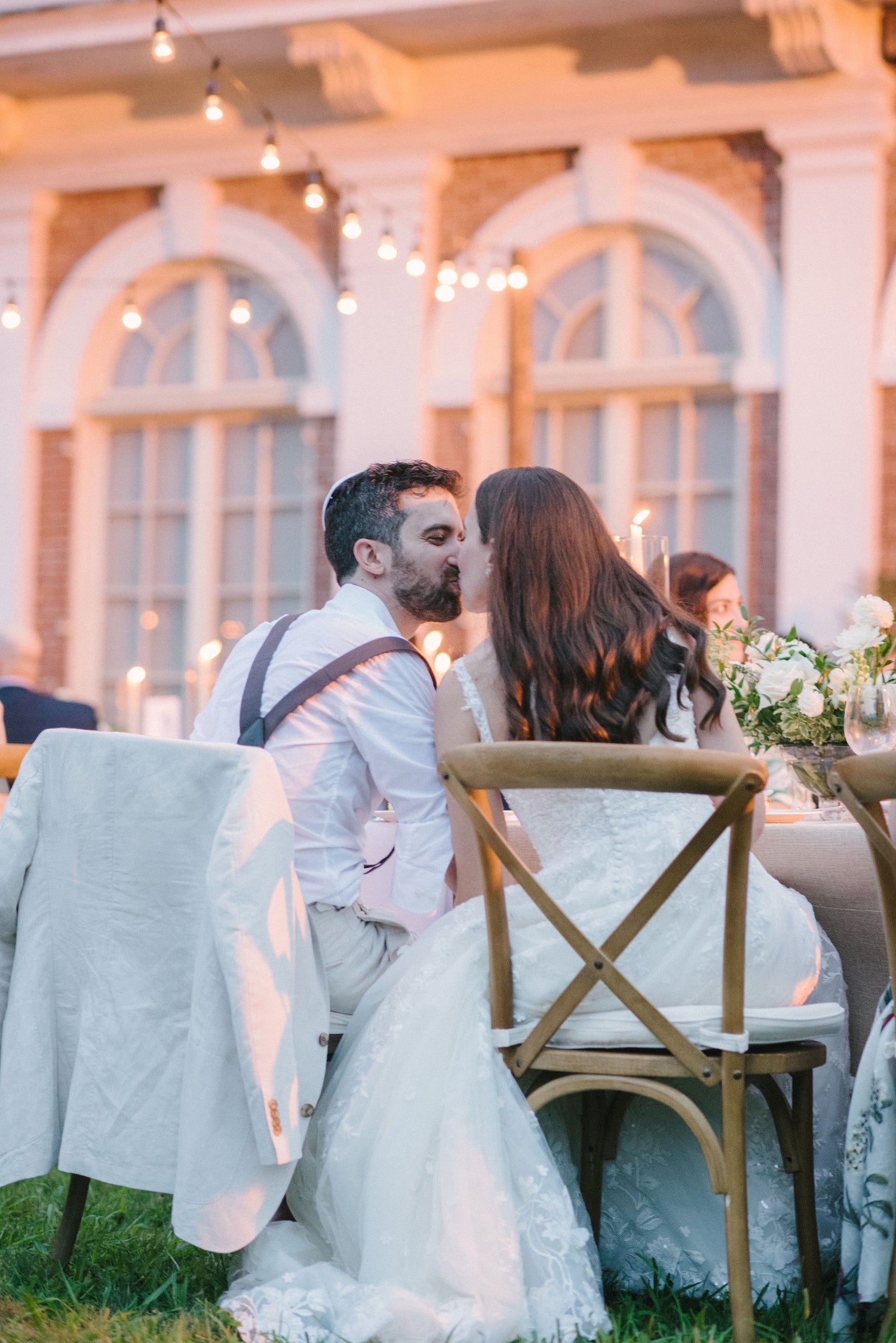 A romantic moment between a bride and groom sharing a kiss during their outdoor wedding reception, surrounded by soft candlelight and floral arrangements.