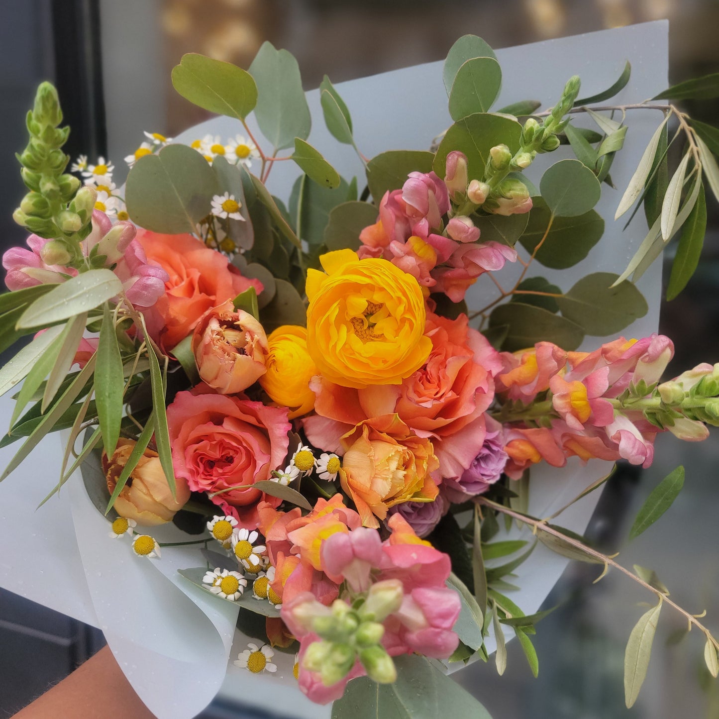 A bright and warm bouquet with yellow ranunculus, peach roses, pink snapdragons, eucalyptus leaves, and white daisies, wrapped in light paper for a fresh look.