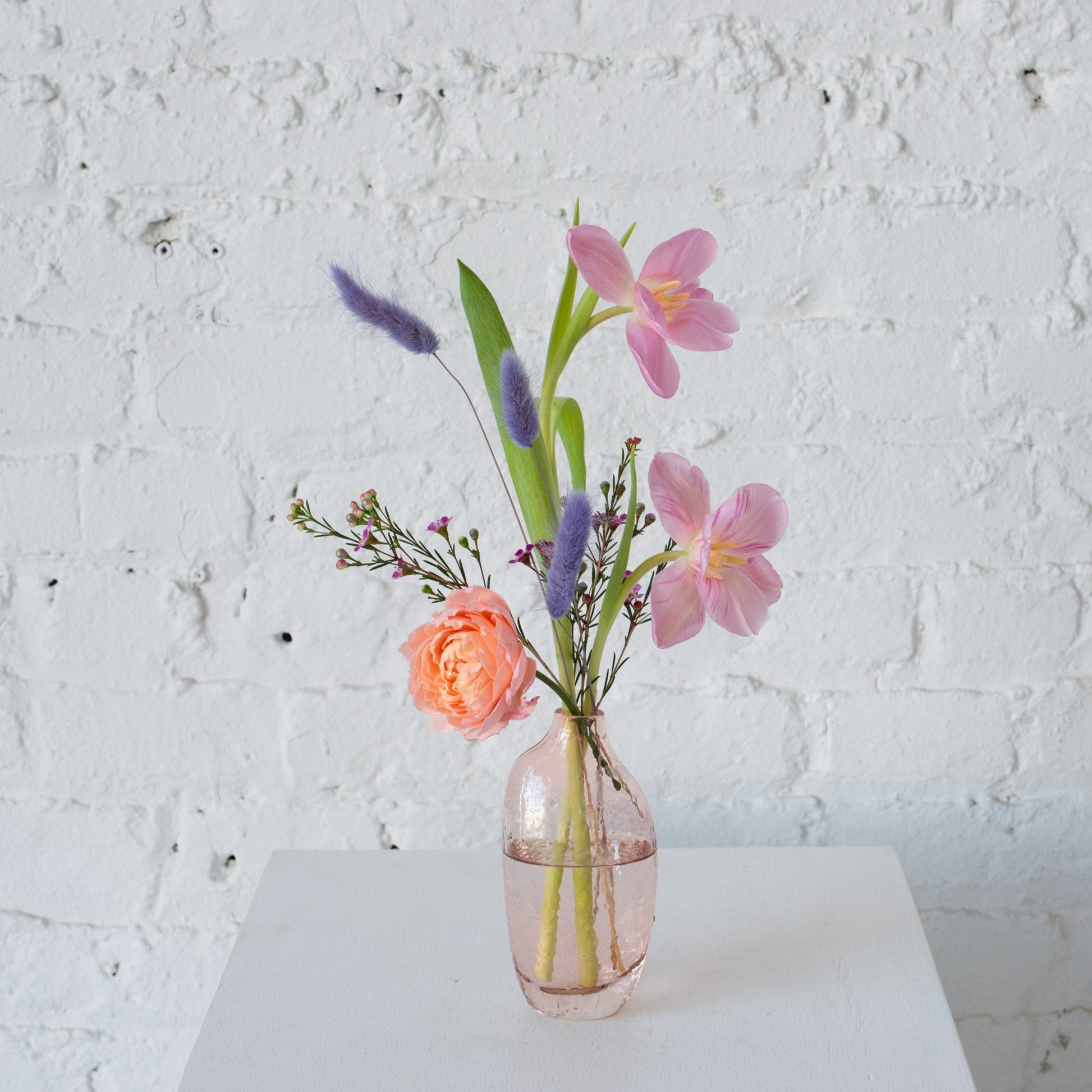 A pastel floral arrangement in a light pink glass bud vase featuring peach garden rose, pink tulips, purple bunny tails, and delicate wax flowers against a white brick background.